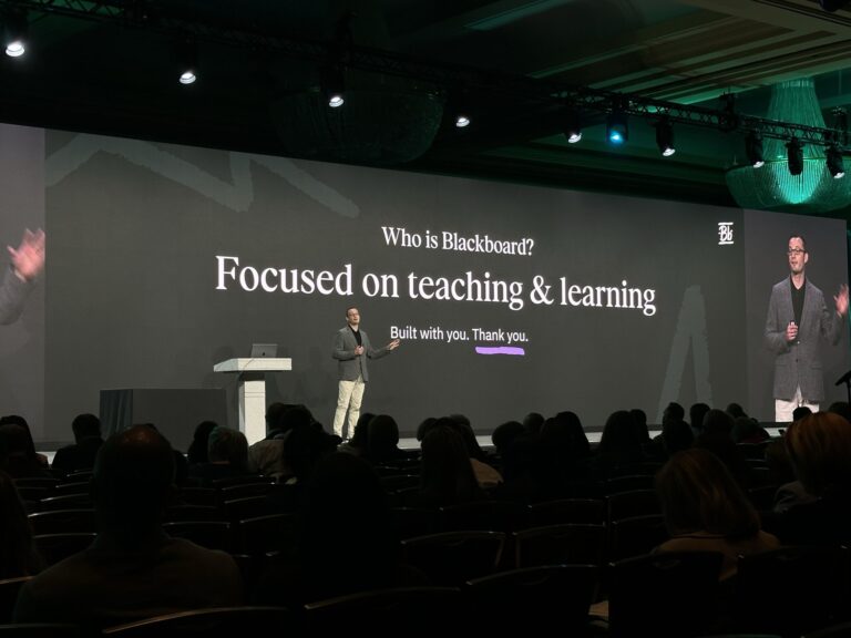 A presenter stands on stage in front of a large screen displaying the text: Who is Blackboard? Focused on teaching & learning. Built with you. Thank you. An audience is seated in the foreground.